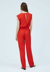 Bretelle rouge sans manches avec un dos en keyhole, une taille élastique et une coupe à jambes larges. Tissu lisse avec une couleur unie, peu de détails.