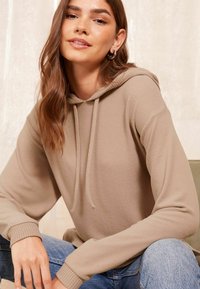 Felpa beige con una texture morbida, dotata di cappuccio con cordino e polsini a costine, abbinata a jeans blu chiaro. Sfondo neutro.