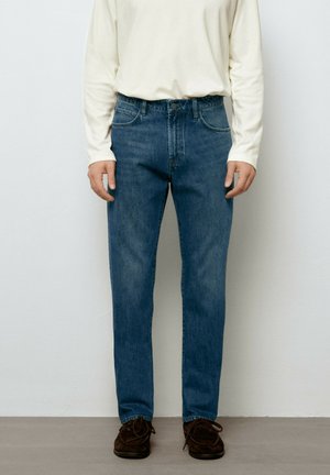 Mann trägt blaue Jeans mit geradem Bein, weißes langärmeliges Hemd und braune Schuhe, steht an einer schlichten Wand.