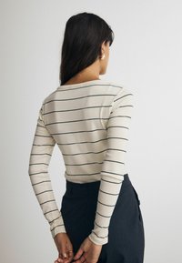 Krämfärgad långärmad topp med svarta horisontella ränder, ribbat mönster, rundad halsringning, matchad med mörka byxor. Slimmad design.