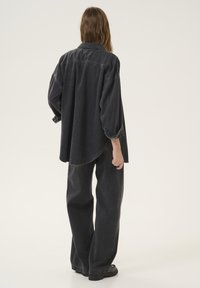 Black denim oversized shirt with a curved hem and rolled sleeves, paired with loose-fitting black denim pants and flat black shoes.