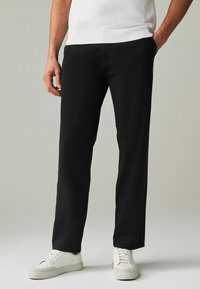 Pantaloni neri in tessuto liscio con vestibilità comoda, dotati di tasche laterali e indossati con sneaker bianche.