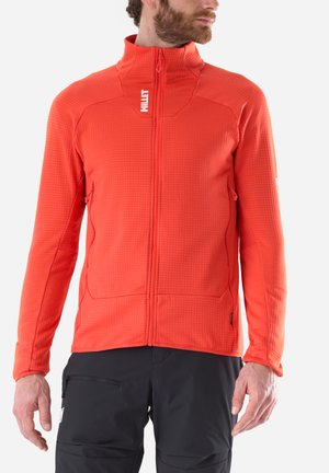 Man wearing bright orange zip-up jacket with high collar and black pants, standing with hands relaxed by sides.
