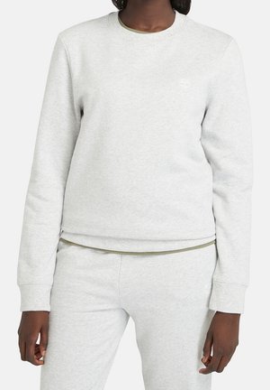 Personne à la peau foncée portant un sweat-shirt gris clair à manches longues et un pantalon de survêtement assorti avec un logo subtil sur la poitrine et la hanche.