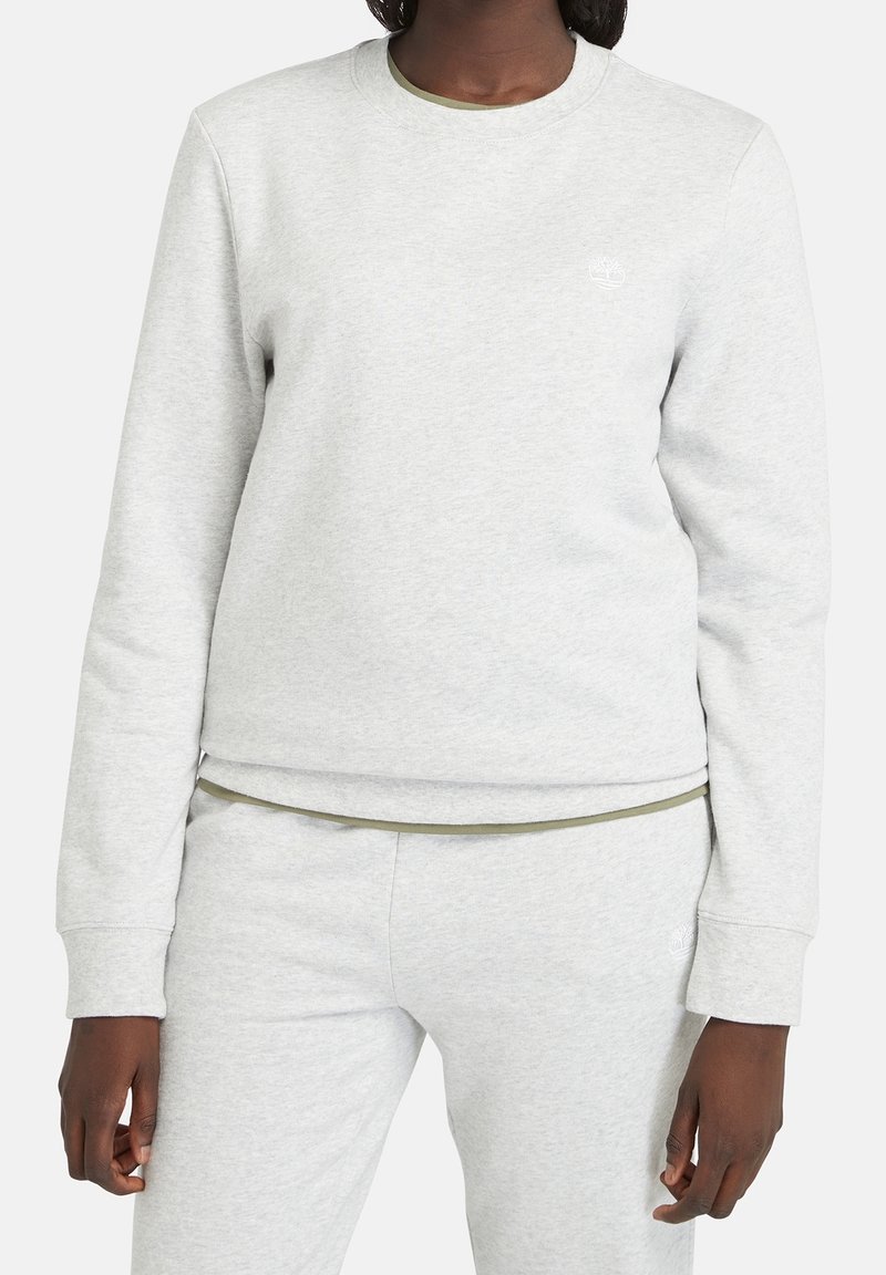 Personne à la peau foncée portant un sweat-shirt gris clair à manches longues et un pantalon de survêtement assorti avec un logo subtil sur la poitrine et la hanche.