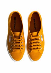 Sneakers in tela gialla con suole in gomma bianca, dotate di sei fori per i lacci, lacci piatti e un'etichetta con logo sul lato. Design in tessuto testurizzato.