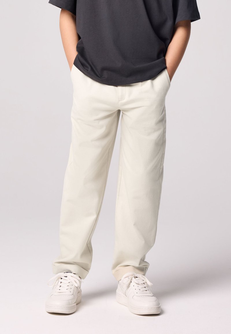 Világos bézs színű egyenes szárú nadrág, pamut anyagból készült. Fehér tornacipővel és sötétszürke pólóval párosítva.