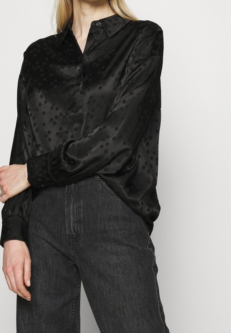 Svart satinblus med ett subtilt prickmönster, krage och knäppning fram. Långa ärmar som avslutas med åtsittande manschetter, ihop med mörkgrå jeans.