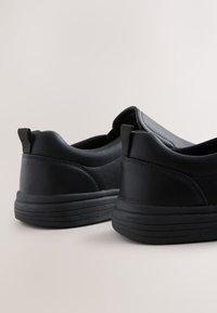 Chaussures slip-on synthétiques noires avec une texture lisse, un design arrondi, des accents minimalistes et une semelle épaisse en caoutchouc présentant des rainures horizontales.