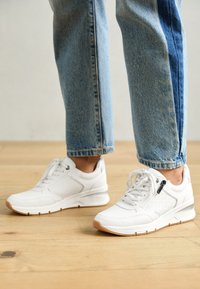 Scarpe da ginnastica bianche con pannelli testurizzati, chiusura con lacci e dettaglio con zip. Suola leggera con un accento in gomma marrone. Indossate con jeans di denim blu.