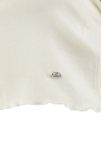Tissu ivoire clair avec une texture lisse, doté d'une petite étiquette ovale en argent portant l'inscription "KEY LARGO" cousue sur le côté. Détail d'ourlet en dentelle.
