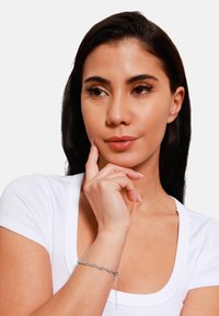 Bracciale in argento con un design delicato, caratterizzato da anelli intrecciati e un piccolo pendente, indossato su una mano su uno sfondo bianco.