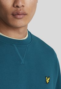 Sudadera de color verde azulado con cuello redondo, costuras visibles y un logotipo amarillo bordado en el lado izquierdo del pecho. Superpuesta a una camiseta verde clara.