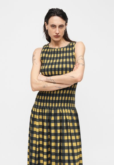 Γυναίκα με σταυρωμένα χέρια, φορώντας μαν sleeveless κίτρινο και μαύρο καρό φόρεμα, στέκεται μπροστά σε ένα απλό ανοιχτό φόντο.