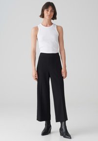 Débardeur côtelé blanc associé à un pantalon noir large taille haute. Le mannequin porte des bottines noires à talons pointus. Design lisse et épuré.