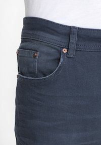Calças de ganga azul-escuro com uma textura suave, apresentando um bolso frontal com rebites de cobre e um design de cós.