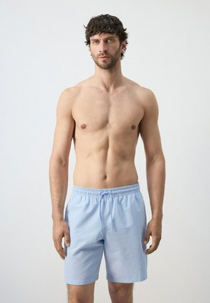 Uomo con capelli scuri che indossa pantaloncini azzurri con coulisse, in piedi contro uno sfondo chiaro semplice, rivolto in avanti con le braccia rilassate.