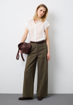 Donna in piedi contro un muro bianco, indossa una camicetta bianca a maniche corte, pantaloni a gamba larga color oliva, cintura marrone, tiene una borsa intrecciata marrone.