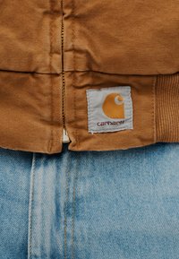 Καφέ μπουφάν Carhartt με ορατό φερμουάρ πάνω από ανοιχτόχρωμο τζιν τζιν, που δείχνει την υφή του υφάσματος και την ετικέτα με το λογότυπο στο τελείωμα του μπουφάν.