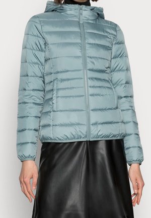 Mujer vestida con chaqueta acolchada con capucha azul claro con cremallera frontal y falda negra, de pie frente a un fondo liso.