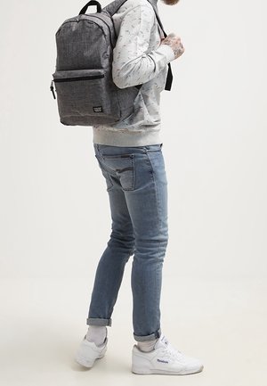Sac à dos en tissu gris avec une poche avant zippée, des bretelles réglables et un design épuré. Porté par une personne portant un sweat à capuche à motifs et un jean.