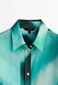 Chemise boutonnée avec un motif dégradé du bleu sarcelle au vert foncé, en tissu lisse, avec un col traditionnel et des coutures visibles.