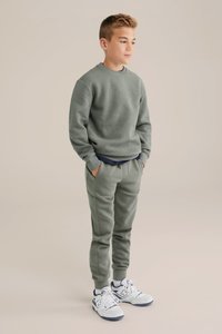 Zelená mikina so vzorom a rebrovými textúrami a zodpovedajúce tepláky, obe vyrobené z mäkkej látky. Biele športové topánky dotvárajú outfit.