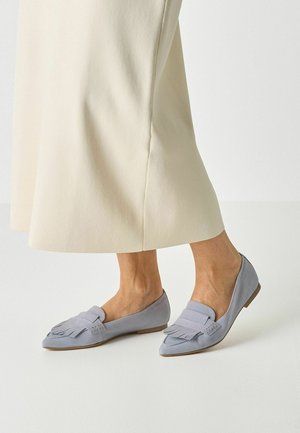 Light gray suede flats with a flat heel, featuring a fringed detail on the toe and a smooth surface. Paired with a long, cream-colored skirt.