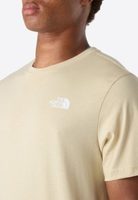 Detailní záběr na osobu nosící béžové tričko s malým bílým logem The North Face na hrudi.