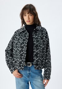 Giacca con stampa leopardata nera e grigia, dotata di colletto, chiusura a bottoni e due tasche laterali, indossata sopra un maglione nero e jeans blu.