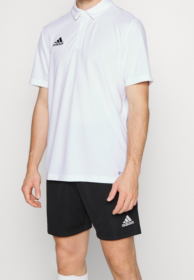 Polo Adidas blanc avec col et trois boutons, accompagné d'un short noir arborant un logo Adidas blanc. Matière en mélange de coton.