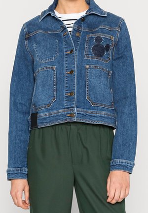 Person trägt eine blaue Jeansjacke mit Knopfverschluss und mehreren Taschen, darunter eine mit einem dunklen Mickey-Mouse-Silhouetten-Aufnäher.