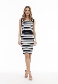 Rochie alb-negru cu model în zigzag, cu o siluetă ajustată și decolteu rotund. Modelul poartă sandale albe cu bretele. Fundalul este alb simplu.