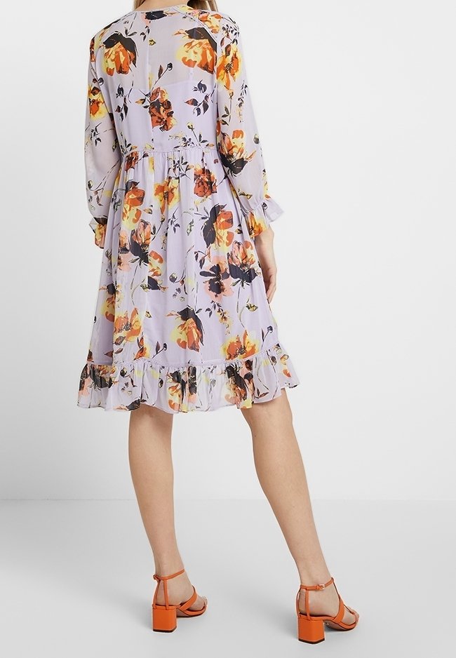 Femme portant une robe lavande à manches longues et longueur genou, imprimée de fleurs orange et noires avec un ourlet à volants, accompagnée de sandales à talons blocs orange.