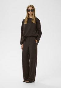 Browna loungewear komplekts ar garām piedurknēm un plašām biksēm. Mīksta auduma, brīva piegrieztne un sānu kabatas. Modelis valkā pārāk lielus melnus saulesbrilles.