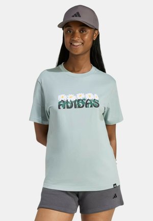 Joven sonriendo, vistiendo una camiseta azul claro de Adidas con logo floral, pantalones cortos grises y gorra gris de Adidas, de pie con las manos detrás de la espalda.