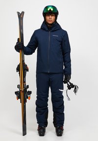 Lyžařský outfit s námořnickou modrou voděodolnou bundou a kalhotami, černými rukavicemi a lyžařskými botami. Drží lyže a lyžařské hůlky s oranžovými akcenty.