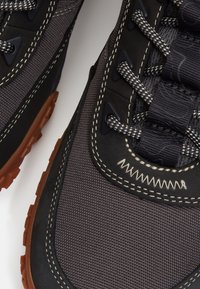 Primo piano di scarpe sportive nere e grigie con cuciture dettagliate, lacci con motivo e suole in gomma marrone testurizzata.