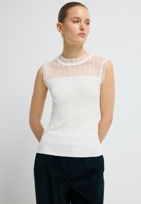 Hvid ærmeløs top med en ribbet tekstur og et gennemsigtigt stribet panel langs halsudskæringen. Har en rund, flæset krave.