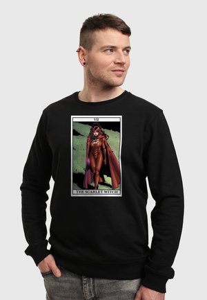 Muž v černé mikině s grafikou tarotové karty Scarlet Witch, kráčející vpřed v červeném oblečení a plášti.