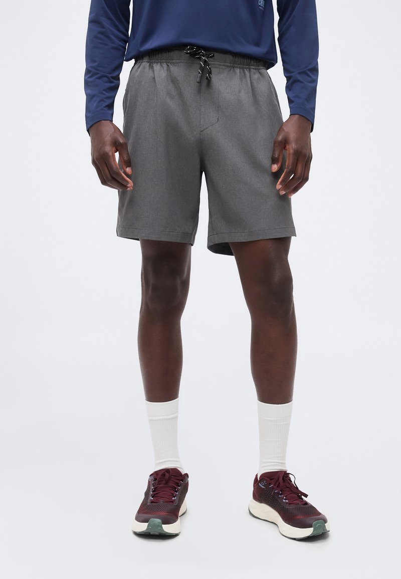 Άνδρας που φορά γκρι σορτς με κορδόνι, λευκές κάλτσες, μπορντό παπούτσια για τρέξιμο και μπλε μακρυμάνικο πουκάμισο, στέκεται σε λευκό φόντο.