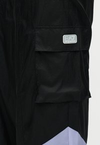 Μαύρη υφασμάτινη τσέπη με καπάκι σε ένδυμα, με ορθογώνιο έμπλαστρο λογότυπου EA7 Emporio Armani σε λευκό και γκρι.