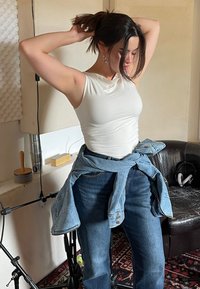 Top bianco aderente senza maniche abbinato a jeans blu a vita alta, giacca di jeans legata in vita, ambiente interno con texture morbide.