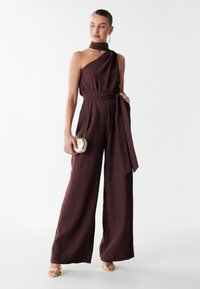 Combinaison plissée bourgogne avec un design à une épaule, taille ceinturée, coupe large et détail de foulard assorti. Modèle tenant une pochette métallique.