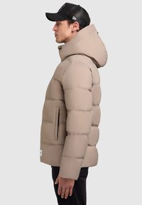Beige pufikabát kapucnival, tűzött mintával és oldalsó zsebekkel. A modellen fekete sapka van, sima, matt felülettel.