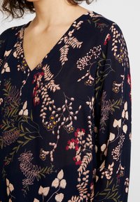 Blouse marine présentant un col en V, des manches longues et une broderie florale complexe dans des tons de rose, rouge et vert sur un tissu texturé.