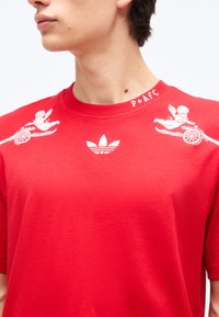 Young person wearing a red T-shirt featuring white winged cherub graphics on shoulders and a central white Adidas trefoil logo.