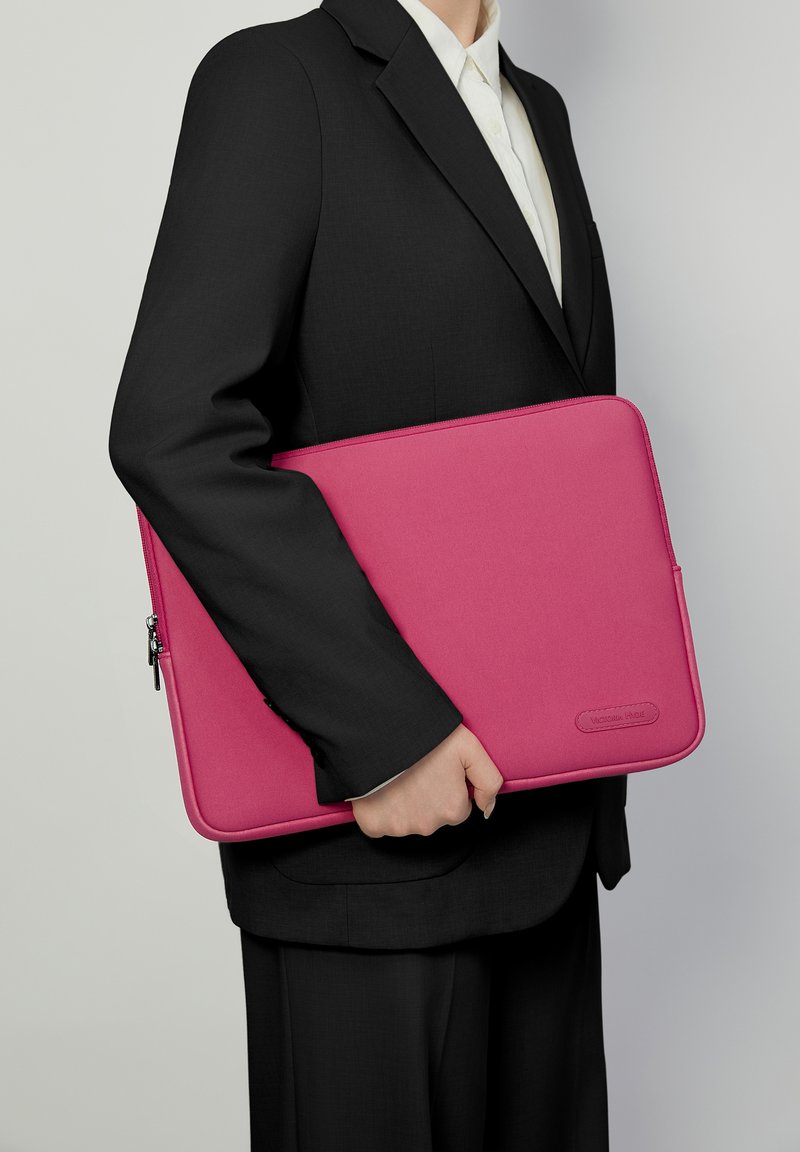 Lys pink laptop sleeve lavet af glat stof, med lynlåslukning og logo-detalje. Holdt mod en sort jakkesæt. Rent, moderne design.