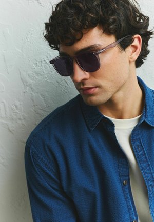 Fiatal férfi göndör sötét hajjal, sötét napszemüveget visel, kék, gombolós ing fehér póló fölött, lefelé néz, világos, texturált fal előtt.