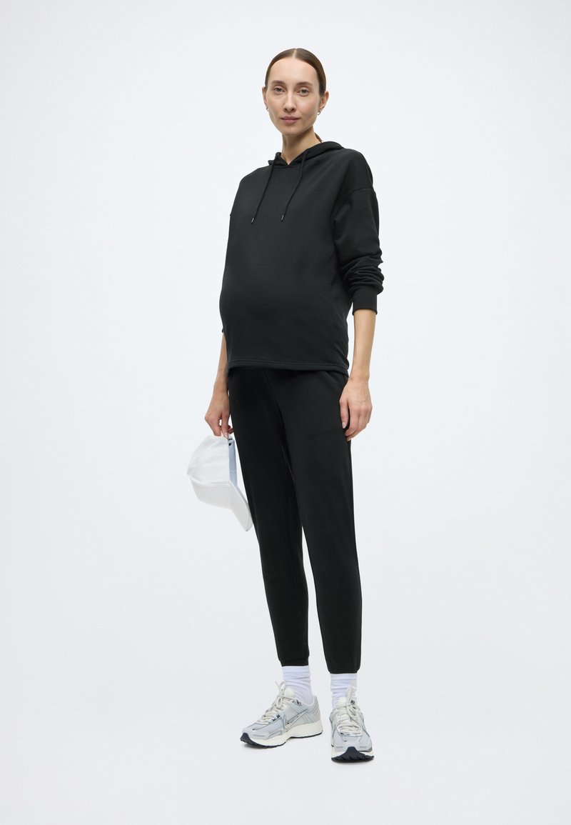 Μαύρο φούτερ με κουκούλα, φαρδιά γραμμή και μπροστινή τσέπη, συνδυασμένο με μαύρο τζόγκερ. Παπούτσια αθλητικά γκρι ανοιχτού χρώματος, κρατώντας ένα λευκό καπέλο στο ένα χέρι.